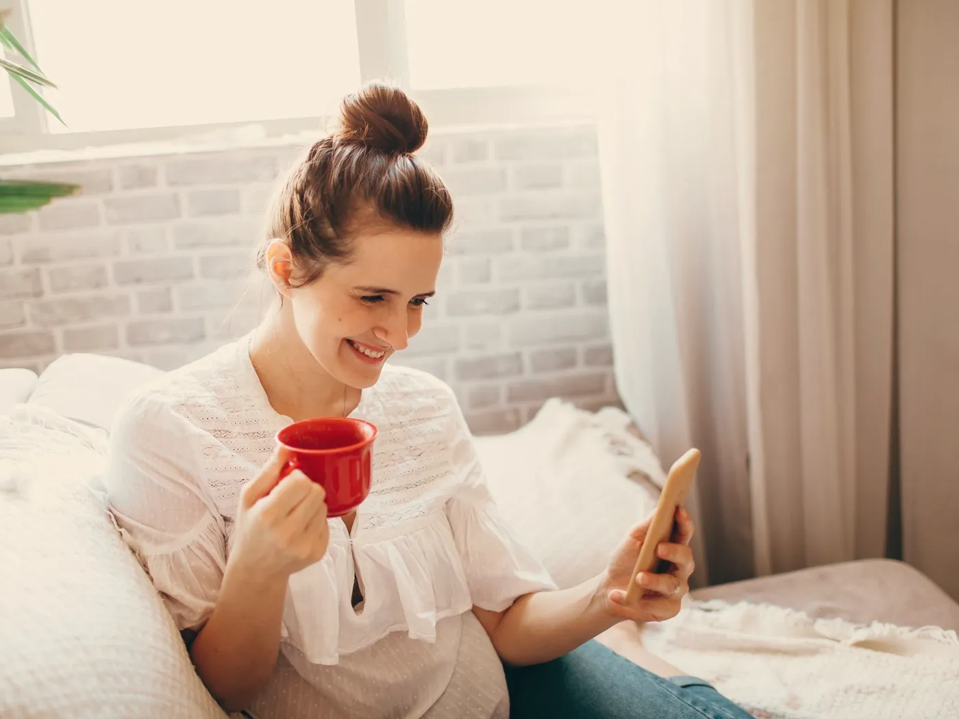 A smiling woman with a cup of coffee looks at her phone on the sofa.